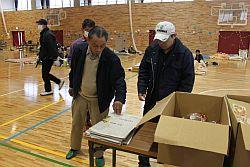 原町第一小学校に避難された男性の方々が机の上にある新聞や物資をみている写真