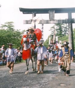 お繰り出しの写真。歴史を感じる石製の鳥居を背に、赤い装いをつけられた馬にまたがった、鎧兜を身に付けている武将をの行進。着物を身にまとった歩兵が武将の周りを囲んでいます。