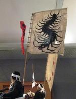 指旗の写真。白地いっぱいに黒下り蜈が描かれており、蚣武将が背にこの旗を掲げた状態で写っています。