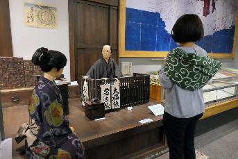 展示室の江戸時代の商家のコーナーの前に風呂敷を背中に背負って立っている筆者