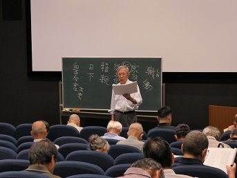板書した黒板の前で資料を読みながら解説する講師とそれを聴く参加者たち