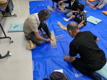 講師がブルーシートに座って竹細工の編み方を実演している