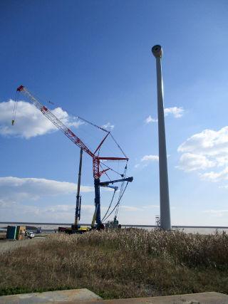 青空の元、据付られた風力発電機のタワーに羽根を取り付ける作業の写真