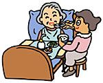 ヘルパーの女性がベットに寝たきりの高齢者女性に食事を与えている画像