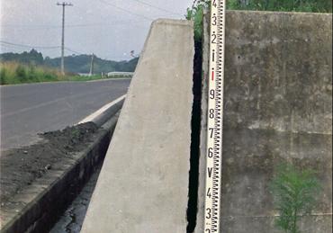 片道1車線の車道右側にある、地場沈下でひびが入った壁に測定メモリが添えられている写真