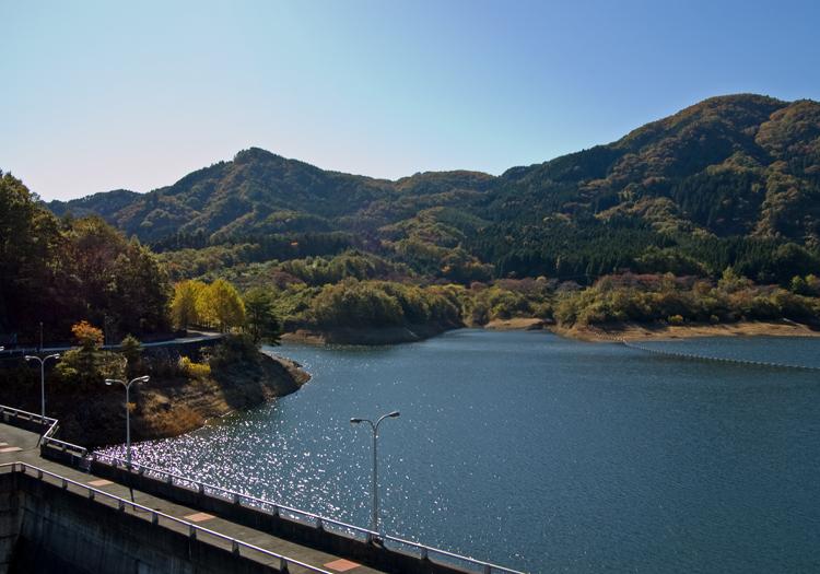 雲一つない青空の下、キラキラ水面が輝いている高の倉ダム貯水池。その奥には山が広がっている写真。