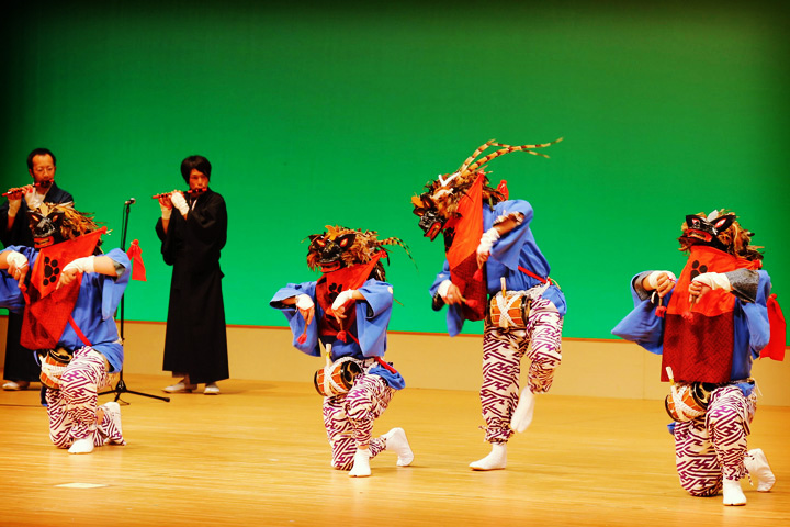 舞台上で獅子舞を披露する4人と横笛を吹く2人の写真