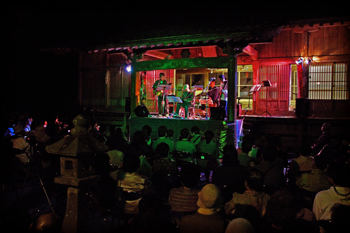 夜の神社の社をステージに演奏するバンドと多くの聴衆の写真