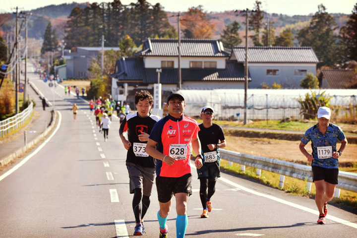 秋、奥の山裾からまっすぐ手前に伸びる道路を連なって走る選手の写真