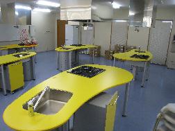 コンロとシンクのある黄色いテーブルが5台ある調理実習室の写真