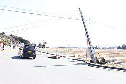 地震の影響で陥没した道路と倒れそうな電柱の横を走る一台の黒い軽自動車の写真