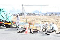 陥没した道路の路肩で下水道の復旧作業をする3人の市職員の写真