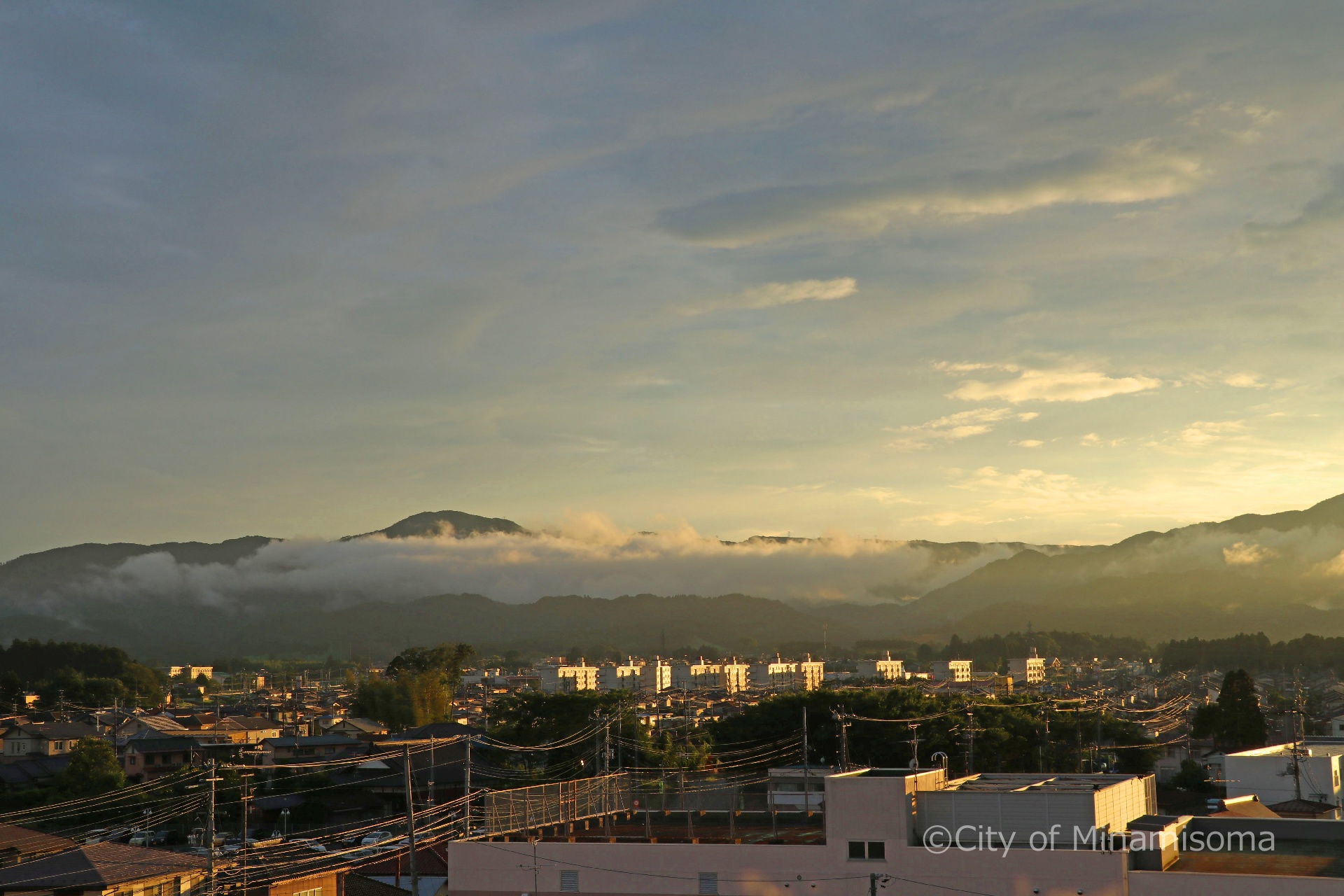 市役所の屋上から見た町の様子。曇り空の隙間から西日が差し、雲がかかった山並みを照らしている。