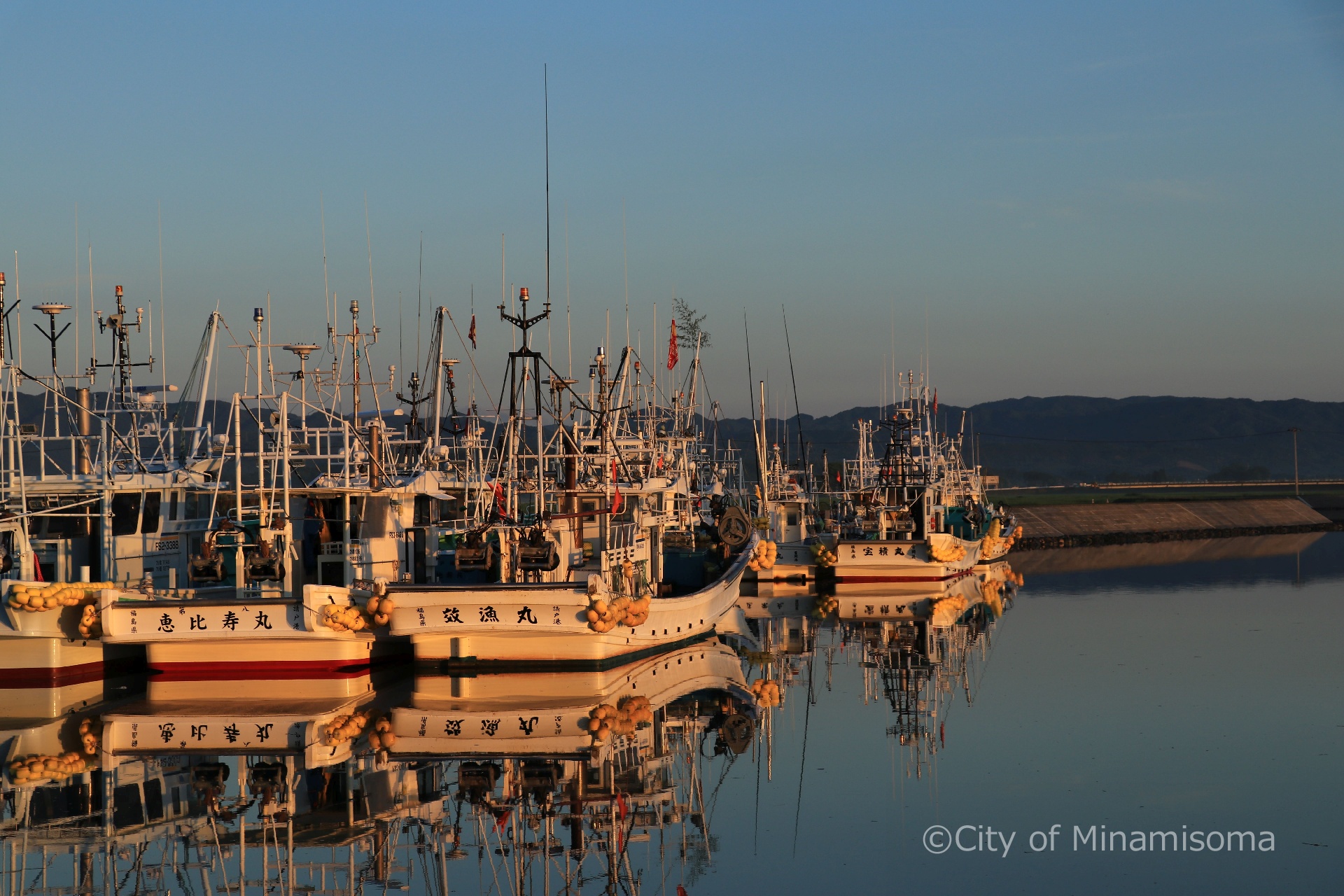 早朝の真野川漁港の様子。鏡のように凪いだ水面に、係留された漁船が映っている。