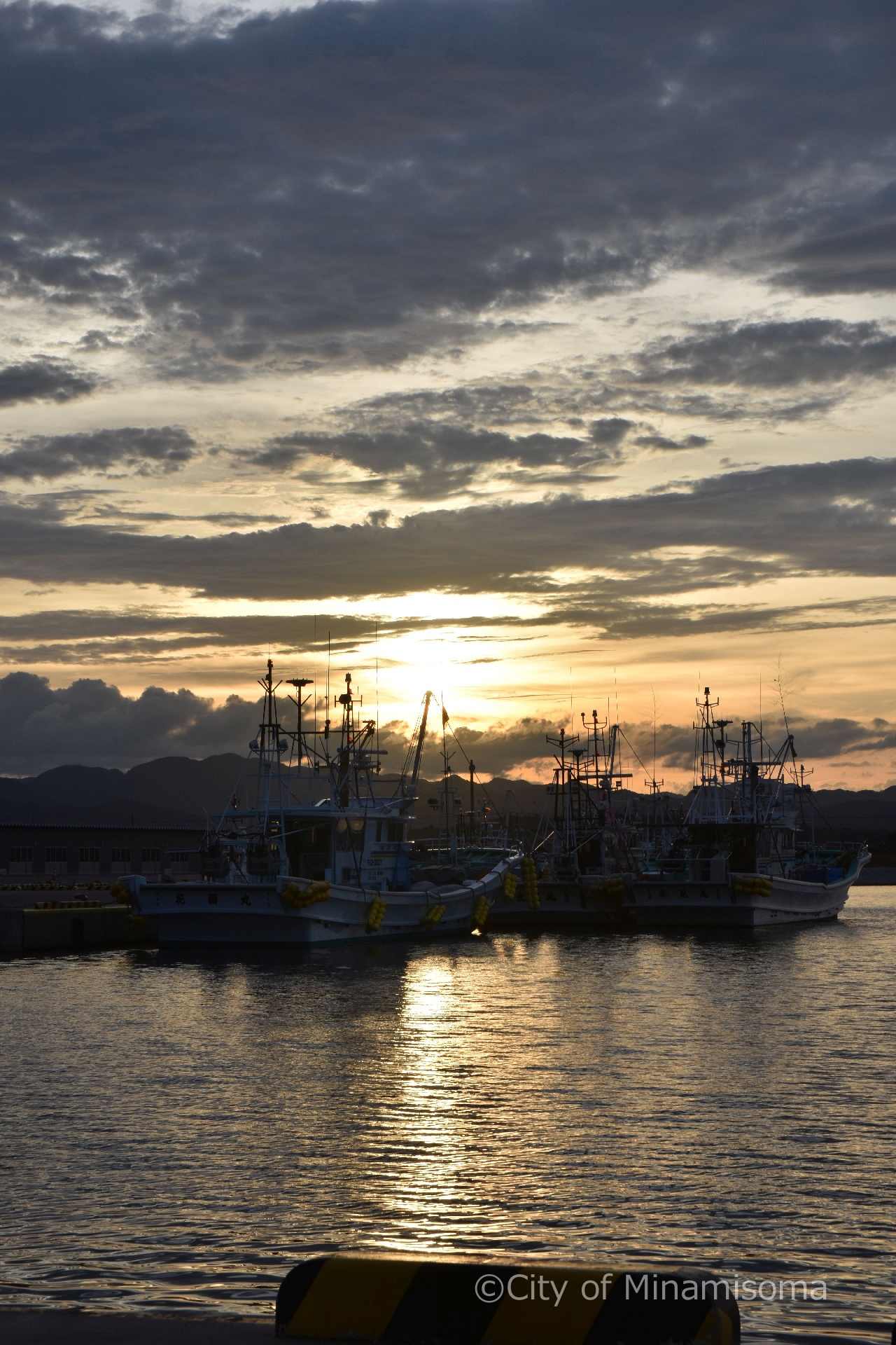 夕方の真野川漁港の様子。灰色の雲の隙間からオレンジ色の太陽が姿を見せ、係留されている漁船のシルエットが浮かび上がっている。
