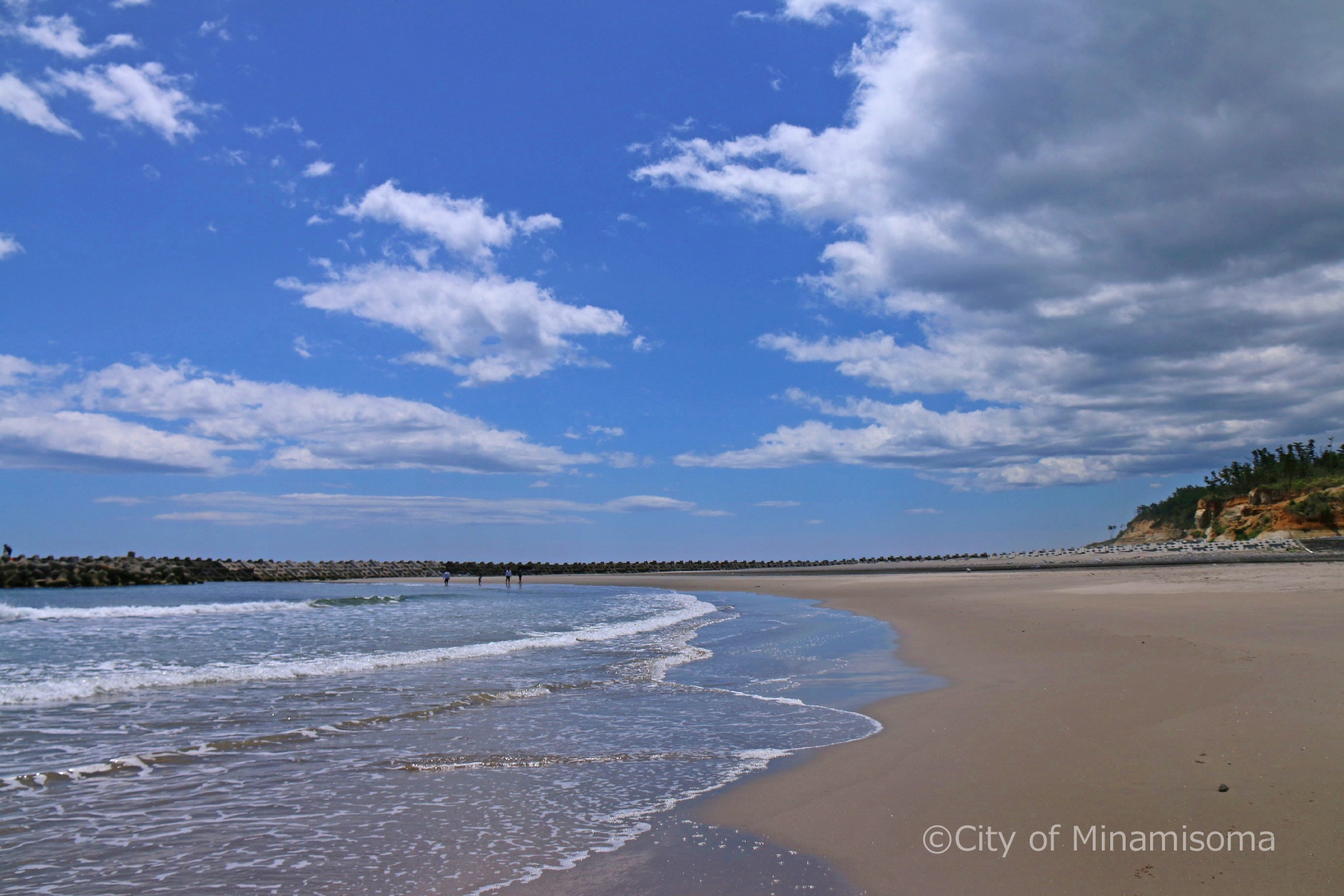 良く晴れた日の北泉海岸の様子。青い空に白い雲が浮かび、遠くの波打ち際を数名の人が歩いている。