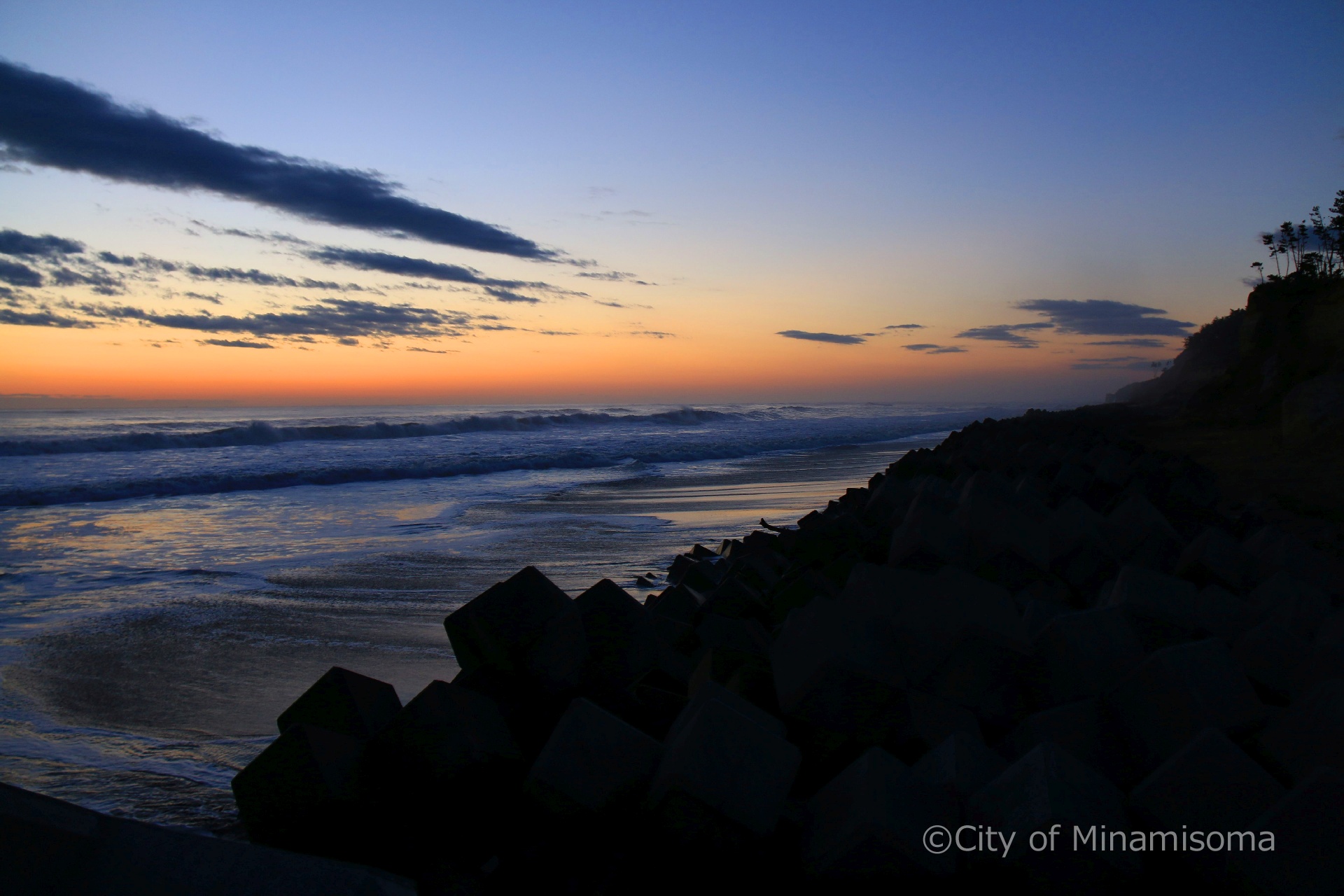 早朝の北泉海岸の様子。青とオレンジのグラデーションの空が、海に映っている。岸壁は黒いシルエットになっている。