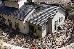 白い二階建て住居の周りを埋め尽く瓦礫の山の写真
