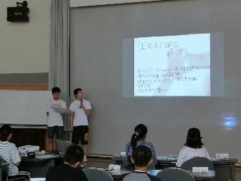 完成した報告会用の発表資料を用いて、男子研修生2人が前に出て発表練習をしている様子が写っている写真