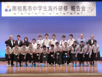 研修生18名、門馬市長、大和田教育長、来賓の方々が報告会の横断幕の前で集合している様子の写真