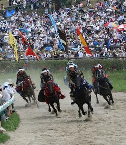 甲冑競馬（雲雀ヶ原）の写真。青や黄色などカラフルな旗を背に掲げた4名の騎馬武者が、右カーブのコーナーを駆け抜ける写真。
