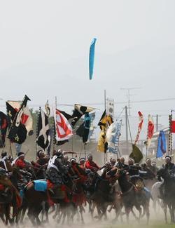 神旗争奪戦（雲雀ヶ原）の写真。街中で、空から落ちてくる水色の神旗を、カラフルな旗を背に掲げた騎馬武者たちが一斉に争奪しあう様子。