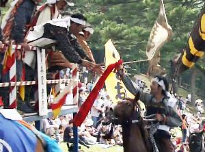 赤い旗を掴んだ騎馬武者が高い場所にいる軍者という野馬追の幹部に旗を手渡しているところ