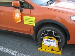 道路上でオレンジ色の車のタイヤ部分にタイヤロック、ミラー部分にミラーズロックがしてあり、黄色い貼り紙が貼ってある写真