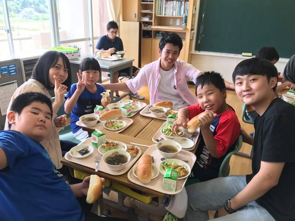 小学生と大学生が給食を一緒に取っている様子の写真